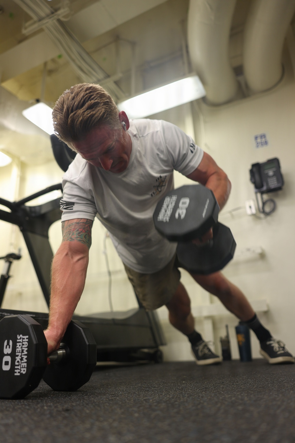 A muscular man with blonde hair and a tattoo on his right forearm is exercising in a gym, performing a dumbbell exercise. He is holding two 30-pound Hammer Strength dumbbells, one on the floor and one raised, suggesting a renegade row or pickup motion. The gym appears to be an institutional or military facility based on the exposed pipes, wall-mounted telephone, and utilitarian interior.