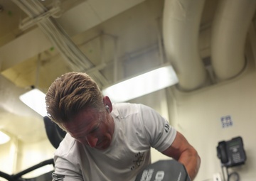 A muscular man with blonde hair and a tattoo on his right forearm is exercising in a gym, performing a dumbbell exercise. He is holding two 30-pound Hammer Strength dumbbells, one on the floor and one raised, suggesting a renegade row or pickup motion. The gym appears to be an institutional or military facility based on the exposed pipes, wall-mounted telephone, and utilitarian interior.