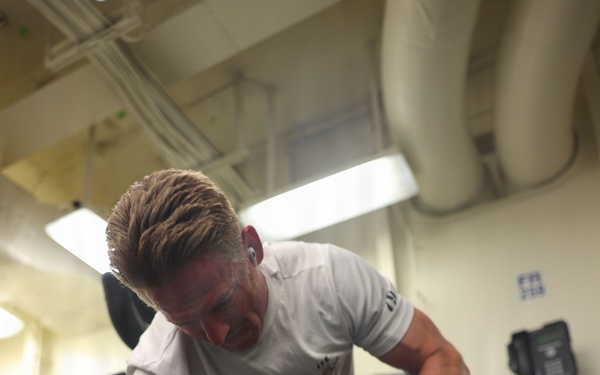 A muscular man with blonde hair and a tattoo on his right forearm is exercising in a gym, performing a dumbbell exercise. He is holding two 30-pound Hammer Strength dumbbells, one on the floor and one raised, suggesting a renegade row or pickup motion. The gym appears to be an institutional or military facility based on the exposed pipes, wall-mounted telephone, and utilitarian interior.