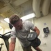 A muscular man with blonde hair and a tattoo on his right forearm is exercising in a gym, performing a dumbbell exercise. He is holding two 30-pound Hammer Strength dumbbells, one on the floor and one raised, suggesting a renegade row or pickup motion. The gym appears to be an institutional or military facility based on the exposed pipes, wall-mounted telephone, and utilitarian interior.