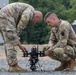 Two U.S. Army soldiers in OCP (Operational Camouflage Pattern) uniforms are inspecting or preparing a small multi-rotor drone on a gravel surface. The drone appears to be a tactical unmanned aerial system (UAS) mounted on landing legs with what appears to be a payload or camera system attached. The setting appears to be a military training or operational area with structures and trees visible in the background.