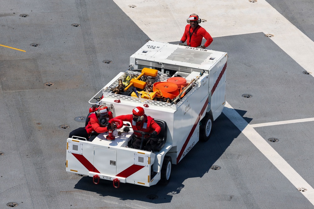 An aerial view of a compact firefighting/crash rescue vehicle on what appears to be an aircraft carrier flight deck. Three personnel dressed in red uniforms and helmets are operating the vehicle, with one standing on top and two seated at the rear. The vehicle is white with red chevron markings and carries firefighting equipment including hoses and tools.
