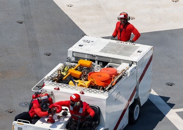 An aerial view of a compact firefighting/crash rescue vehicle on what appears to be an aircraft carrier flight deck. Three personnel dressed in red uniforms and helmets are operating the vehicle, with one standing on top and two seated at the rear. The vehicle is white with red chevron markings and carries firefighting equipment including hoses and tools.