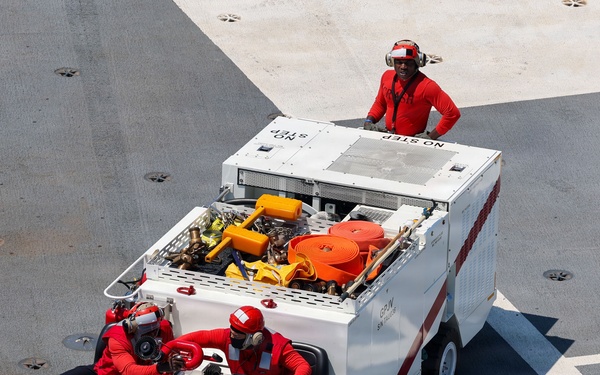An aerial view of a compact firefighting/crash rescue vehicle on what appears to be an aircraft carrier flight deck. Three personnel dressed in red uniforms and helmets are operating the vehicle, with one standing on top and two seated at the rear. The vehicle is white with red chevron markings and carries firefighting equipment including hoses and tools.
