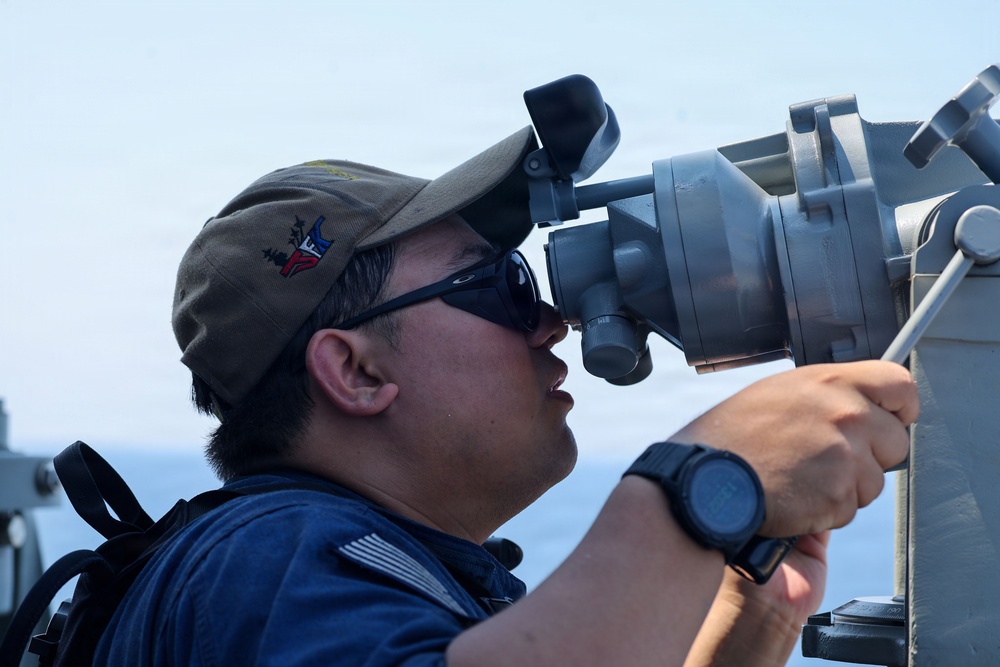 A military service member is using a large optical sighting or navigation instrument aboard a naval vessel at sea. The individual is wearing a dark navy uniform, Oakley sunglasses, and a military-style cap with an emblem. The background shows open ocean and clear sky conditions.