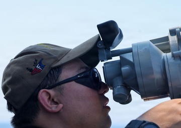 A military service member is using a large optical sighting or navigation instrument aboard a naval vessel at sea. The individual is wearing a dark navy uniform, Oakley sunglasses, and a military-style cap with an emblem. The background shows open ocean and clear sky conditions.