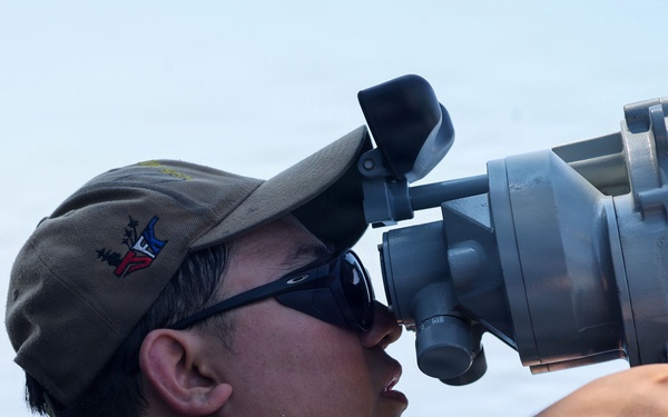 A military service member is using a large optical sighting or navigation instrument aboard a naval vessel at sea. The individual is wearing a dark navy uniform, Oakley sunglasses, and a military-style cap with an emblem. The background shows open ocean and clear sky conditions.