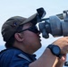 A military service member is using a large optical sighting or navigation instrument aboard a naval vessel at sea. The individual is wearing a dark navy uniform, Oakley sunglasses, and a military-style cap with an emblem. The background shows open ocean and clear sky conditions.