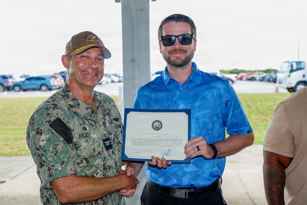 A U.S. Navy officer in camouflage NWU (Navy Working Uniform) with the name tape 'PRATT' and captain's eagle insignia is shaking hands with a civilian man in a blue polo shirt. The civilian is holding a Navy certificate or award document featuring an official seal. The ceremony appears to be taking place outdoors in a covered area near a parking lot.
