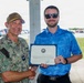 A U.S. Navy officer in camouflage NWU (Navy Working Uniform) with the name tape 'PRATT' and captain's eagle insignia is shaking hands with a civilian man in a blue polo shirt. The civilian is holding a Navy certificate or award document featuring an official seal. The ceremony appears to be taking place outdoors in a covered area near a parking lot.
