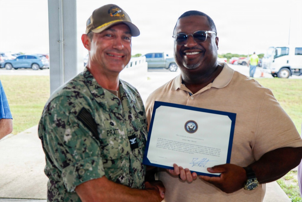 Two men are posing together while holding a formal certificate or commendation document bearing a U.S. Navy seal. The man on the left is wearing U.S. Navy digital camouflage (NWU) uniform with a name tape reading 'PRATT' and a military cap, while the man on the right is in civilian attire holding the framed certificate. The setting appears to be an outdoor covered area at what looks like a military installation.