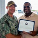 Two men are posing together while holding a formal certificate or commendation document bearing a U.S. Navy seal. The man on the left is wearing U.S. Navy digital camouflage (NWU) uniform with a name tape reading 'PRATT' and a military cap, while the man on the right is in civilian attire holding the framed certificate. The setting appears to be an outdoor covered area at what looks like a military installation.