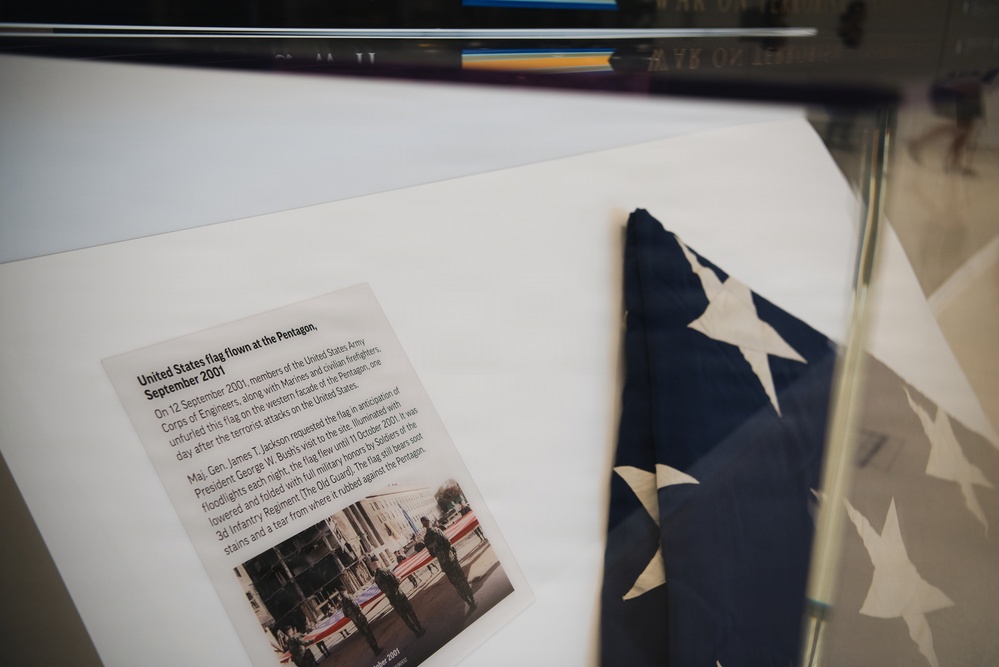 A museum or memorial display case containing a folded United States flag and an accompanying placard. The exhibit identifies this as the U.S. flag flown at the Pentagon in September 2001, one day after the September 11 terrorist attacks. The flag was unfurled by members of the U.S. Army Corps of Engineers, Marines, and civilian firefighters on September 12, 2001.