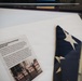 A museum or memorial display case containing a folded United States flag and an accompanying placard. The exhibit identifies this as the U.S. flag flown at the Pentagon in September 2001, one day after the September 11 terrorist attacks. The flag was unfurled by members of the U.S. Army Corps of Engineers, Marines, and civilian firefighters on September 12, 2001.
