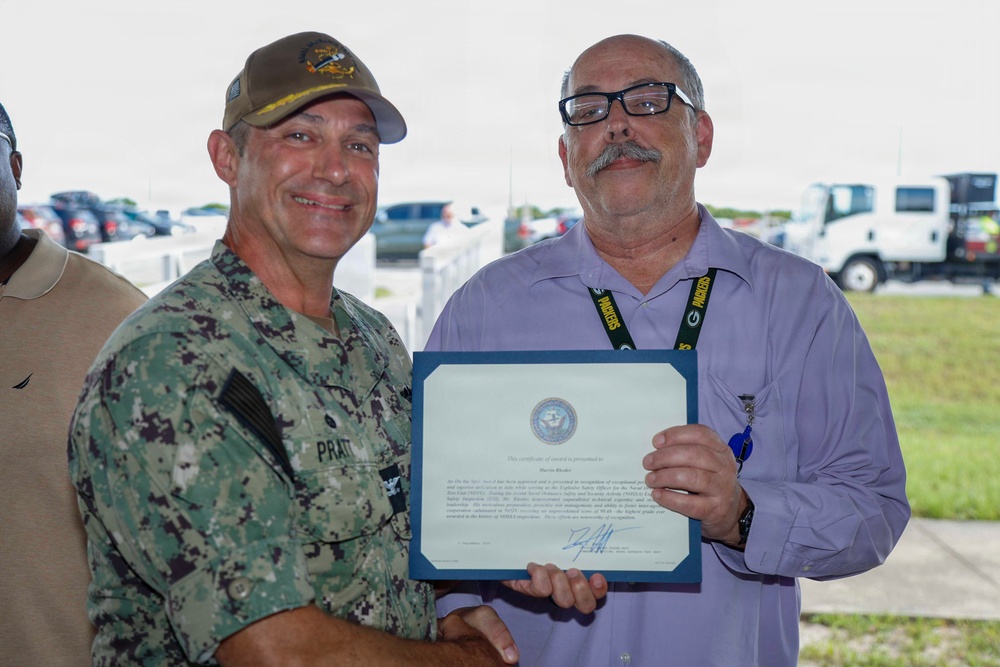 Two men are posing together while holding a certificate of award or recognition. The man on the left is wearing a U.S. Navy digital camouflage uniform with a name tape reading 'PRATT' and a military cap, while the man on the right is in civilian attire wearing glasses and a lanyard. The ceremony appears to be taking place outdoors in a parking or staging area.