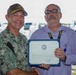 Two men are posing together while holding a certificate of award or recognition. The man on the left is wearing a U.S. Navy digital camouflage uniform with a name tape reading 'PRATT' and a military cap, while the man on the right is in civilian attire wearing glasses and a lanyard. The ceremony appears to be taking place outdoors in a parking or staging area.