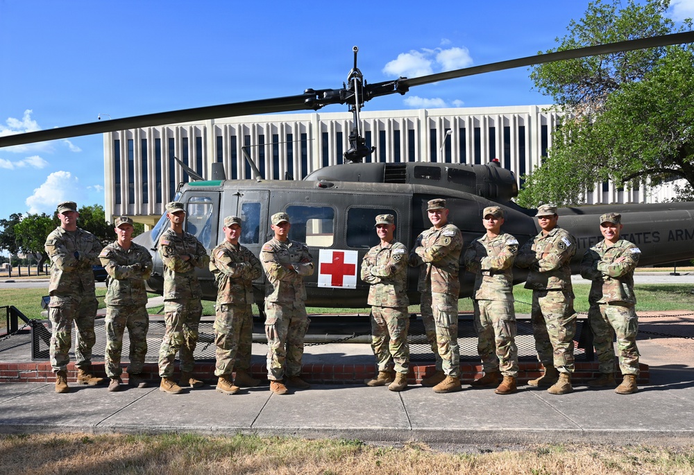 A group of nine U.S. Army soldiers in MultiCam/OCP camouflage uniforms pose in front of a static display UH-1 'Huey' medevac helicopter marked with a Red Cross symbol. The helicopter bears 'UNITED STATES ARMY' lettering on its tail boom and is mounted on a raised platform, suggesting it is a memorial or static display piece. A large institutional building is visible in the background, consistent with a military installation.