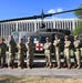 A group of nine U.S. Army soldiers in MultiCam/OCP camouflage uniforms pose in front of a static display UH-1 'Huey' medevac helicopter marked with a Red Cross symbol. The helicopter bears 'UNITED STATES ARMY' lettering on its tail boom and is mounted on a raised platform, suggesting it is a memorial or static display piece. A large institutional building is visible in the background, consistent with a military installation.