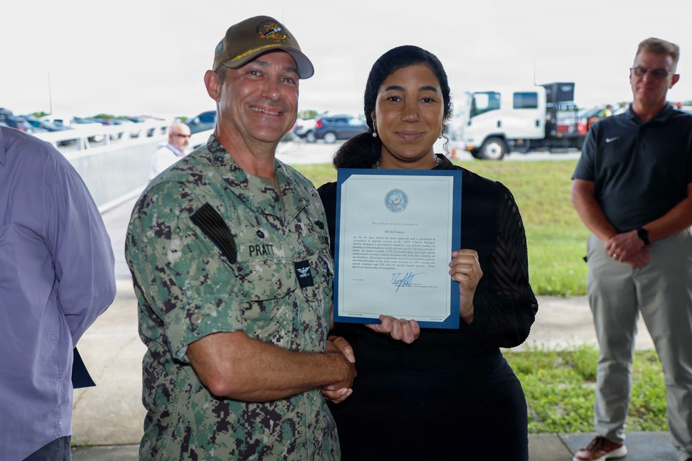 A U.S. Navy officer identified by name tape as 'PRATT' poses with a civilian woman who is holding a framed certificate or commendation document. The ceremony appears to be taking place outdoors at what looks like a military installation or base. The officer is wearing Navy Working Uniform (NWU) camouflage and the two individuals are shaking hands for the photo.
