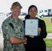 A U.S. Navy officer identified by name tape as 'PRATT' poses with a civilian woman who is holding a framed certificate or commendation document. The ceremony appears to be taking place outdoors at what looks like a military installation or base. The officer is wearing Navy Working Uniform (NWU) camouflage and the two individuals are shaking hands for the photo.