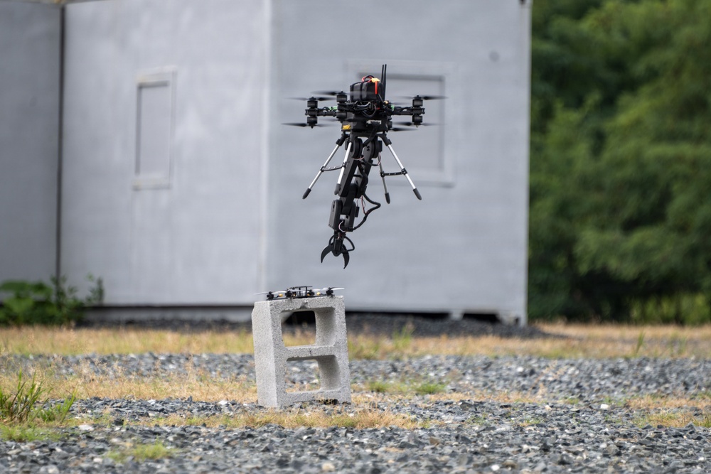 A large multi-rotor drone equipped with a robotic claw/gripper arm is hovering above two stacked concrete cinder blocks on a gravel surface. The drone appears to be in the process of picking up or releasing the cinder blocks, suggesting a payload manipulation or delivery test. The scene takes place outdoors near a gray modular structure, likely a controlled testing environment.