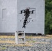 A large multi-rotor drone equipped with a robotic claw/gripper arm is hovering above two stacked concrete cinder blocks on a gravel surface. The drone appears to be in the process of picking up or releasing the cinder blocks, suggesting a payload manipulation or delivery test. The scene takes place outdoors near a gray modular structure, likely a controlled testing environment.