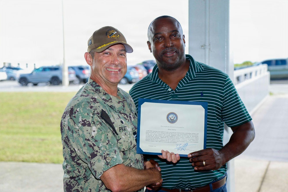 Two men are posing together while holding a framed certificate or award document. The man on the left is wearing U.S. Navy camouflage (NWU) uniform with a name tape reading 'PRATT' and a military ball cap, while the man on the right is in civilian attire (green striped polo shirt). The setting appears to be an outdoor covered area at a military installation, with a parking lot visible in the background.