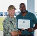 Two men are posing together while holding a framed certificate or award document. The man on the left is wearing U.S. Navy camouflage (NWU) uniform with a name tape reading 'PRATT' and a military ball cap, while the man on the right is in civilian attire (green striped polo shirt). The setting appears to be an outdoor covered area at a military installation, with a parking lot visible in the background.