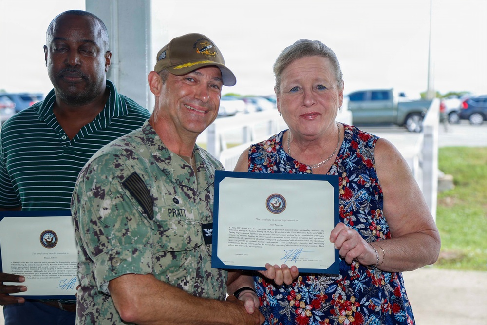A U.S. Navy service member in digital camouflage uniform, identified by name tape as 'PRATT', presents a certificate of award to a woman in a floral dress. A second man in a green striped polo shirt stands in the background holding a similar certificate. The setting appears to be an outdoor covered area near a parking lot, likely on a military installation.