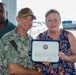 A U.S. Navy service member in digital camouflage uniform, identified by name tape as 'PRATT', presents a certificate of award to a woman in a floral dress. A second man in a green striped polo shirt stands in the background holding a similar certificate. The setting appears to be an outdoor covered area near a parking lot, likely on a military installation.