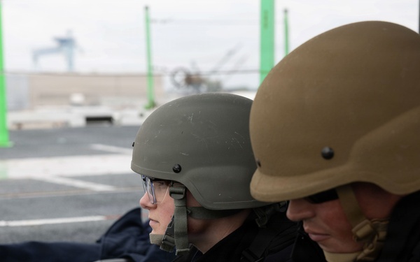 Two individuals wearing military-style ballistic helmets and protective eyewear are positioned behind a belt-fed machine gun, likely during a training exercise. Both are dressed in dark tactical jackets and appear to be at a firing range or aboard a military vessel. An ammunition box with linked rounds is visible in the foreground.