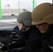 Two individuals wearing military-style ballistic helmets and protective eyewear are positioned behind a belt-fed machine gun, likely during a training exercise. Both are dressed in dark tactical jackets and appear to be at a firing range or aboard a military vessel. An ammunition box with linked rounds is visible in the foreground.