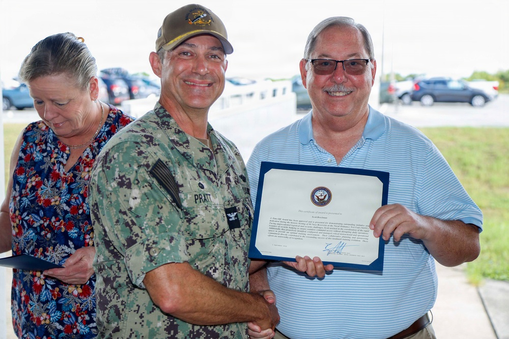 A U.S. Navy officer identified by name tape as 'PRATT' presents a certificate of award to a civilian man in a blue striped polo shirt. The certificate bears a Navy seal and appears to be a formal recognition award. A woman in a floral dress stands in the background, appearing to read from a document.
