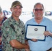 A U.S. Navy officer identified by name tape as 'PRATT' presents a certificate of award to a civilian man in a blue striped polo shirt. The certificate bears a Navy seal and appears to be a formal recognition award. A woman in a floral dress stands in the background, appearing to read from a document.