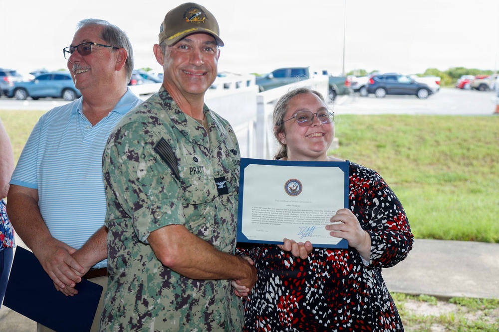A U.S. Navy officer in digital camouflage uniform (NWU) with the name tape 'PRATT' is presenting a certificate to a young woman wearing glasses and a red and white floral dress. The certificate appears to be an official Navy recognition document displayed in a blue folder. A civilian man in a light blue polo shirt stands in the background, smiling.