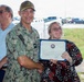 A U.S. Navy officer in digital camouflage uniform (NWU) with the name tape 'PRATT' is presenting a certificate to a young woman wearing glasses and a red and white floral dress. The certificate appears to be an official Navy recognition document displayed in a blue folder. A civilian man in a light blue polo shirt stands in the background, smiling.