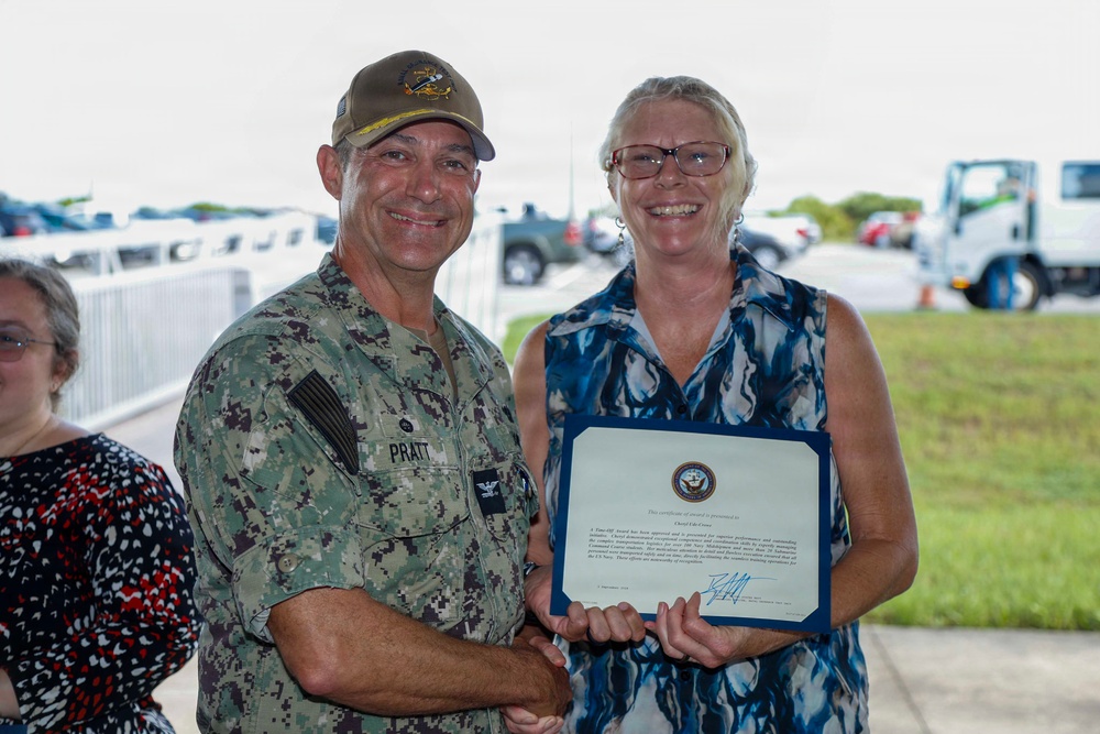A U.S. Navy officer in digital camouflage uniform is presenting a certificate of award to a civilian woman outdoors. Both individuals are smiling and holding the framed certificate together. The setting appears to be a military installation based on the vehicles and infrastructure visible in the background.