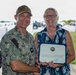 A U.S. Navy officer in digital camouflage uniform is presenting a certificate of award to a civilian woman outdoors. Both individuals are smiling and holding the framed certificate together. The setting appears to be a military installation based on the vehicles and infrastructure visible in the background.