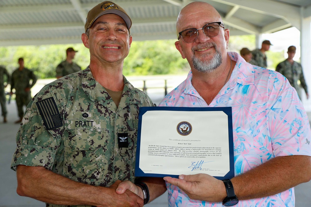 A U.S. Navy officer with the name tag 'PRATT' is presenting a certificate of award to a civilian man in a floral shirt. Both men are shaking hands and smiling while holding the certificate together. The ceremony appears to be taking place at an outdoor covered area with other military personnel visible in the background.