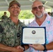 A U.S. Navy officer with the name tag 'PRATT' is presenting a certificate of award to a civilian man in a floral shirt. Both men are shaking hands and smiling while holding the certificate together. The ceremony appears to be taking place at an outdoor covered area with other military personnel visible in the background.