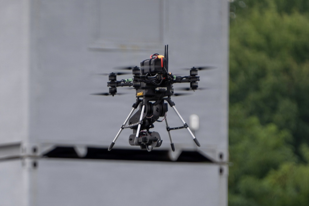 A professional-grade multirotor drone (hexacopter or octocopter) is captured mid-flight against a blurred gray structure background with green trees visible to the right. The drone is equipped with a gimbal-mounted camera payload suspended beneath its frame via a landing gear assembly. The image is sharp and well-focused on the drone, suggesting a telephoto lens was used.