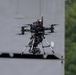 A professional-grade multirotor drone (hexacopter or octocopter) is captured mid-flight against a blurred gray structure background with green trees visible to the right. The drone is equipped with a gimbal-mounted camera payload suspended beneath its frame via a landing gear assembly. The image is sharp and well-focused on the drone, suggesting a telephoto lens was used.