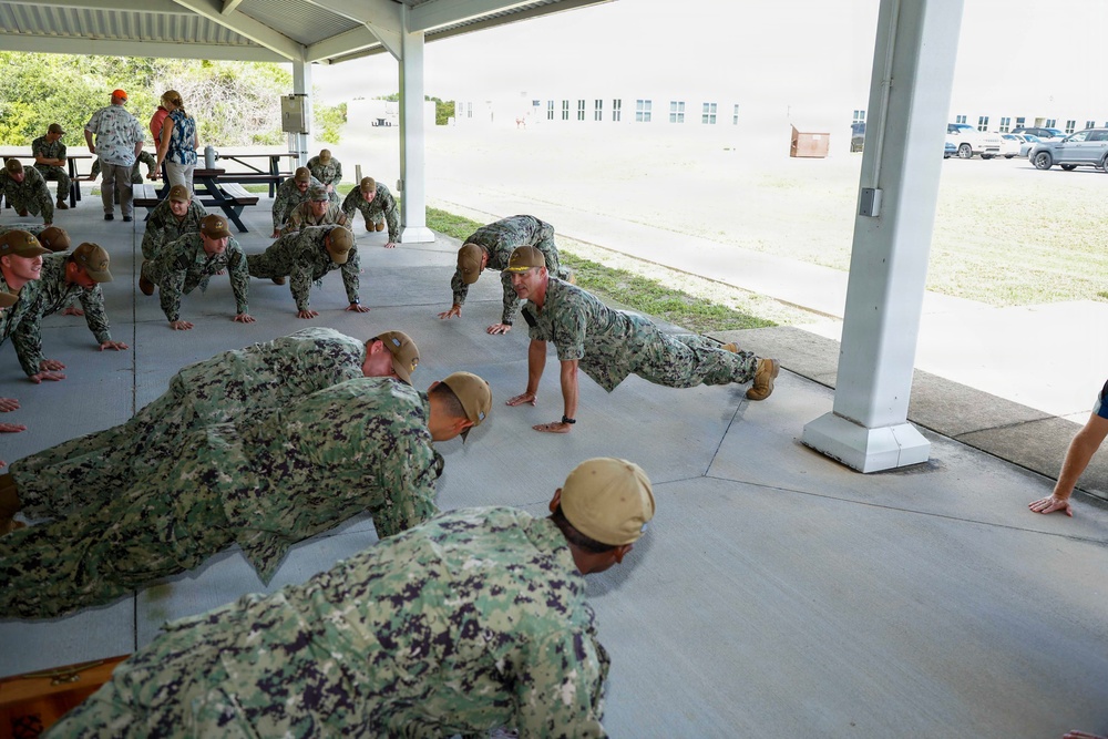 A group of military personnel in Navy Working Uniform (NWU) camouflage are performing push-ups under a covered outdoor pavilion. One individual, distinguished by a yellow headband, appears to be in a leadership or instructor role, demonstrating proper form. The setting appears to be a military base with institutional buildings and vehicles visible in the background.