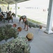 A group of military personnel in Navy Working Uniform (NWU) camouflage are performing push-ups under a covered outdoor pavilion. One individual, distinguished by a yellow headband, appears to be in a leadership or instructor role, demonstrating proper form. The setting appears to be a military base with institutional buildings and vehicles visible in the background.