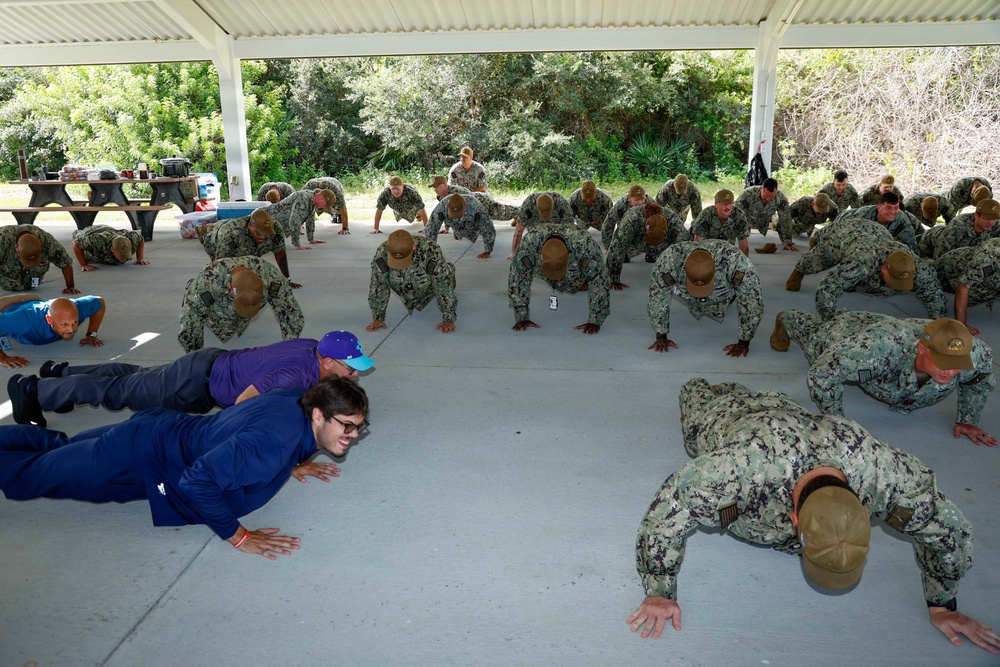 A large group of military personnel in Navy Working Uniform (NWU) digital camouflage are performing push-ups under an outdoor covered pavilion. Two civilians in casual clothing are also participating alongside the uniformed service members. The setting appears to be a military base or outdoor recreational area with trees visible in the background.