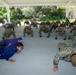 A large group of military personnel in Navy Working Uniform (NWU) digital camouflage are performing push-ups under an outdoor covered pavilion. Two civilians in casual clothing are also participating alongside the uniformed service members. The setting appears to be a military base or outdoor recreational area with trees visible in the background.