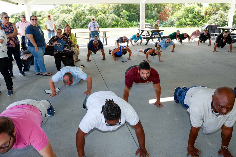 A group of people are performing push-ups on a concrete surface under an outdoor covered pavilion. Several observers stand and sit along the sides watching the exercise activity. The setting appears to be a park or recreational area with picnic tables and green trees visible in the background.