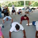 A group of people are performing push-ups on a concrete surface under an outdoor covered pavilion. Several observers stand and sit along the sides watching the exercise activity. The setting appears to be a park or recreational area with picnic tables and green trees visible in the background.