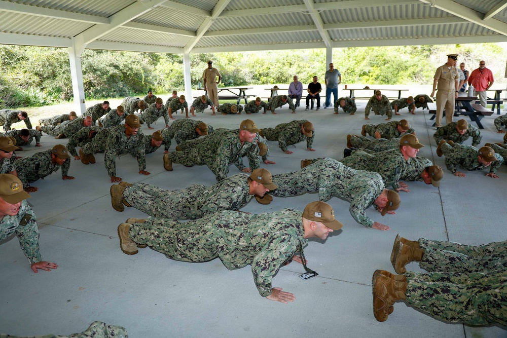 A large group of military personnel in U.S. Navy Working Uniform (NWU Type III camouflage) are performing push-ups in unison under an open-air pavilion. Several observers, including what appears to be a senior officer in khaki uniform, watch from the perimeter. The setting appears to be an outdoor recreational or training area with picnic tables and natural vegetation in the background.