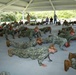 A large group of military personnel in U.S. Navy Working Uniform (NWU Type III camouflage) are performing push-ups in unison under an open-air pavilion. Several observers, including what appears to be a senior officer in khaki uniform, watch from the perimeter. The setting appears to be an outdoor recreational or training area with picnic tables and natural vegetation in the background.